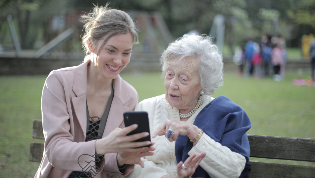 A younger woman calmly and kindly explains something to an elderly woman while they sit on a bench in a park; the image shows a warm, trusting conversation.
