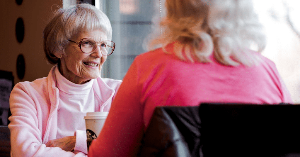 A younger woman sitting at eye level, holding hands and communicating kindly with an elderly woman in a home setting.