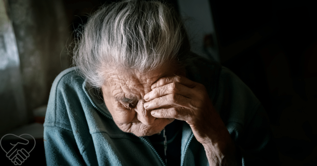 An elderly woman sits alone in a dimly lit room, her face partially obscured by her hands as she looks down, conveying a sense of withdrawal and lack of engagement.