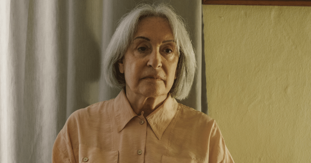 An elderly woman sitting alone in her room, looking thoughtfully into the distance by a window.
