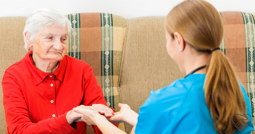 A heartwarming scene of an elderly woman with dementia sitting at home, looking at her caregiver. The caregiver is sitting opposite her with their back to the camera, gently holding the elderly woman's hands in a supportive and comforting gesture.