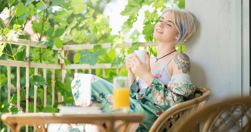 A young woman sitting on a sunny balcony, eyes closed with a peaceful smile, holding a warm mug of tea. The background shows soft plants and gentle light, conveying a sense of calm and much-needed rest.