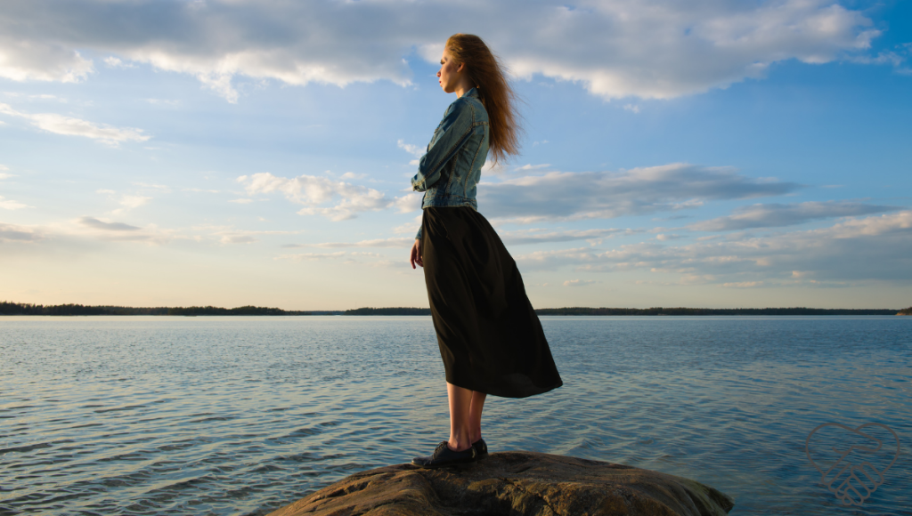 Side view of a woman standing confidently on a cliff by the sea, wind blowing her hair as she looks towards the horizon with trust and resilience.