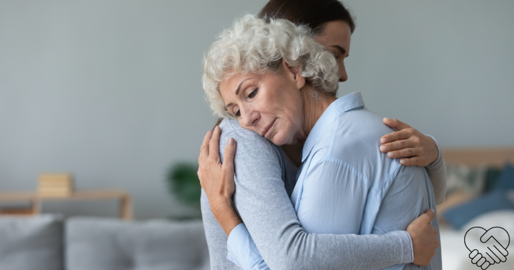 A younger woman comforting an elderly woman with a warm hug at home, representing emotional support for dementia caregivers.