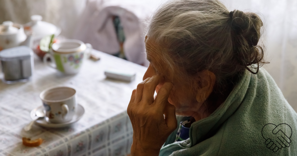 An elderly woman sitting alone at a wooden table, seen from behind, holding her head in her hands with an untouched cup of tea in front of her, illustrating the frustration of swallowing difficulties in dementia.