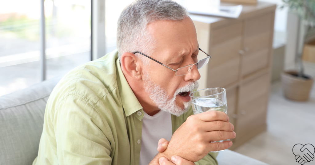 n elderly man at home, sitting at a table, holding a glass of water with both hands, looking focused and slightly overwhelmed as he tries to drink.