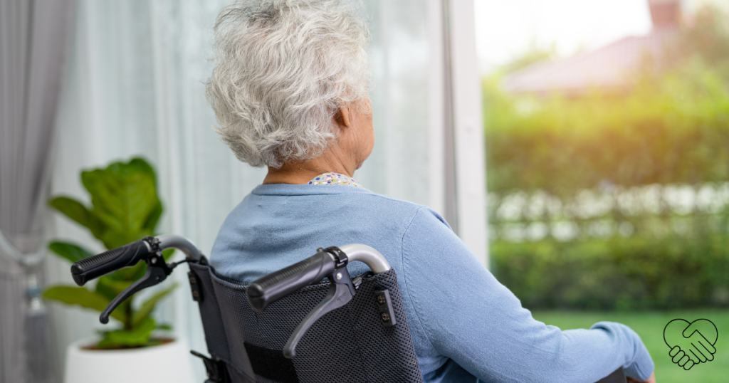 An elderly loved one with dementia sitting in a wheelchair in a bright room, looking out an open window, symbolizing peace and safety at home.