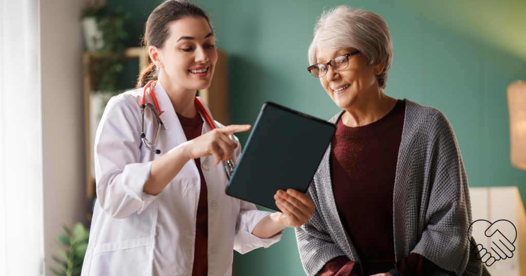 A young, dark-haired female doctor smiling while presenting digital biomarker results on a tablet to an elderly woman in a modern medical office. The woman is smiling with relief upon hearing the positive news.