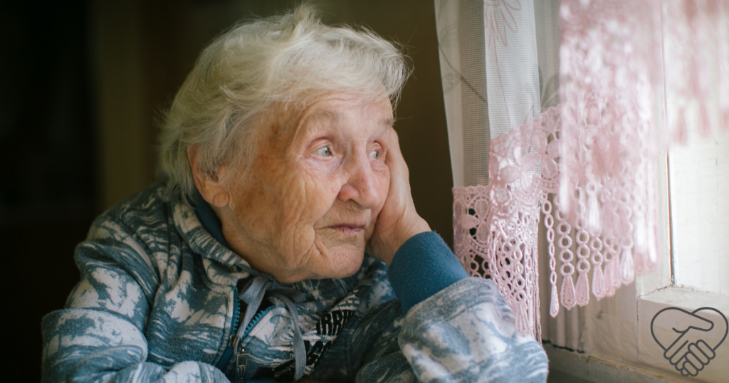 An elderly woman with dementia sitting at home in her room, looking out the window with a blank, distant stare.
