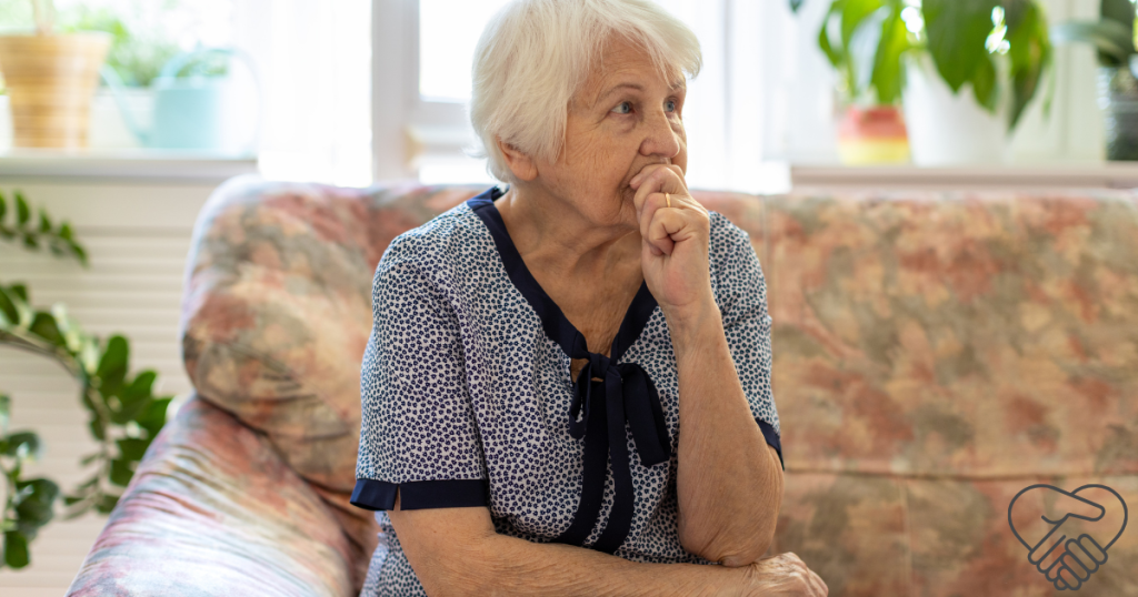 An elderly woman sitting on a sofa in a living room, looking towards a corner with an anxious and frightened expression, illustrating a moment of dementia-related hallucination.