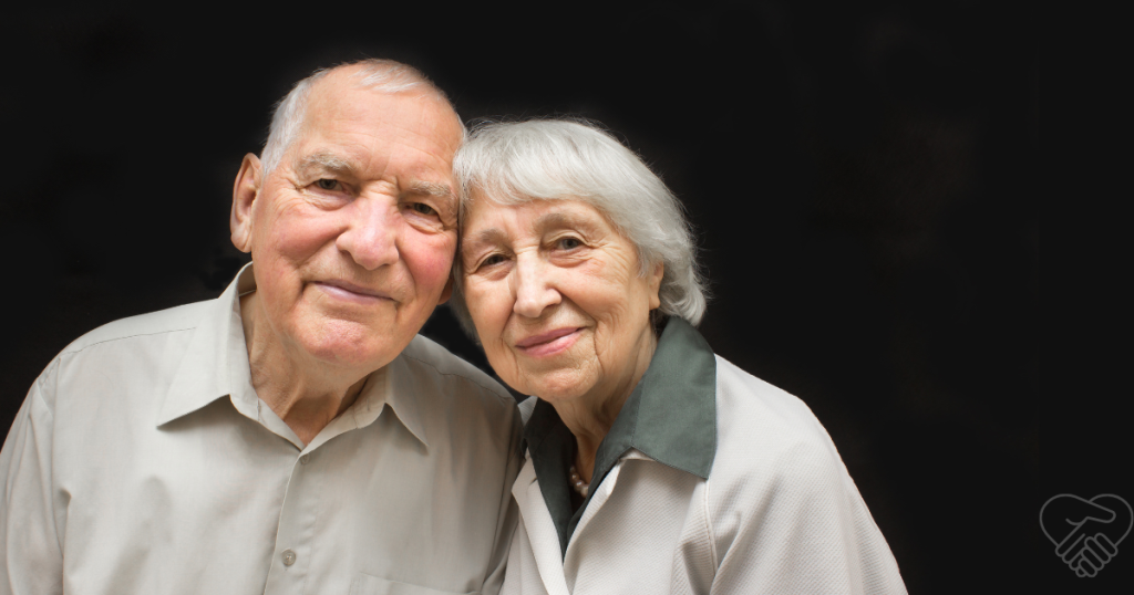 An elderly couple leaning their heads together against a neutral background, showing a quiet, warm moment of connection and resilience while living with dementia.