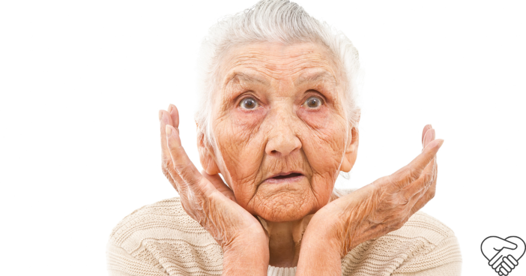 High-angle medium shot of an elderly woman standing in an empty space, holding her hands to her face with a deeply confused and bewildered expression.