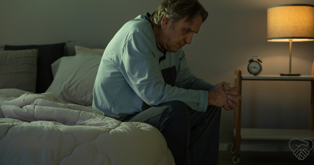 An elderly man with dementia sitting on the edge of his bed at night in a dark room, unable to sleep.