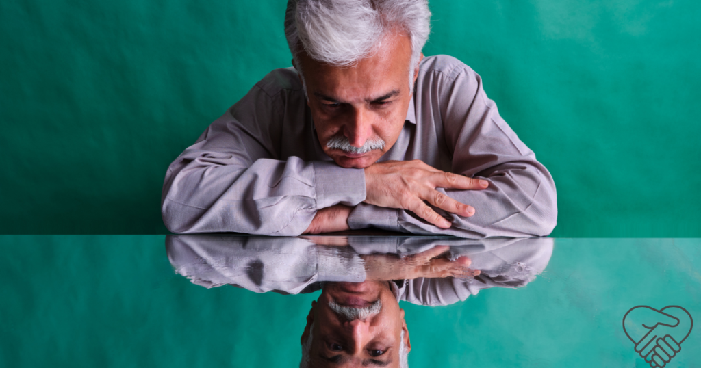 Frontotemporal Dementia – What you need to know as a caregiver A man sitting at a table looking at his own reflection on the polished surface. The reflection appears distant and distorted, symbolizing the loss of self in dementia.