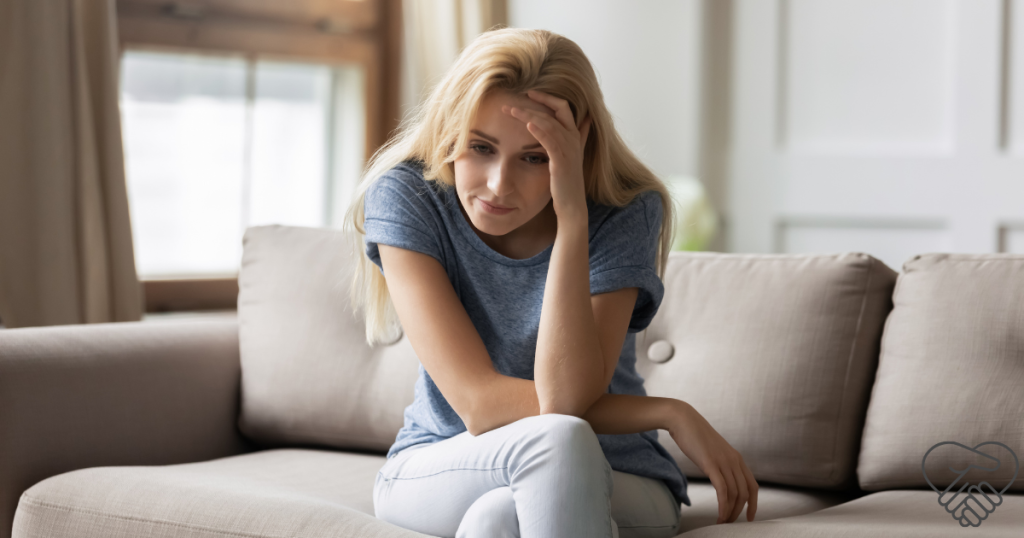 Caregiver Burnout in Dementia Care: Signs and Symptoms to Watch For An exhausted woman sitting on a sofa in a living room, leaning forward and holding her head with one hand. The image illustrates the physical and emotional burden of caregiver burnout.